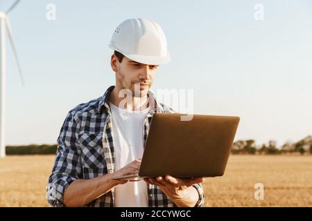 An electrical engineer standing on a field with windmills, using laptop computer, checking on wind turbine energy production Stock Photo