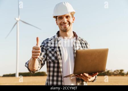 An electrical engineer standing on a field with windmills, using laptop computer, thumbs up Stock Photo
