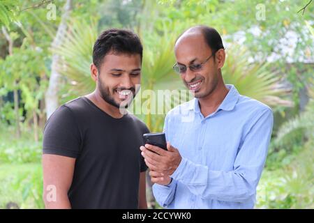 Casual Asian father showing news feed on the mobile phone to his son outside, both smiling while watching Stock Photo