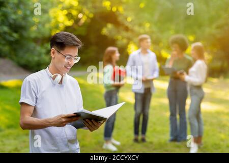 Happy Chinese Male Student Resting At Campus After Classes, Holding Workbook Stock Photo