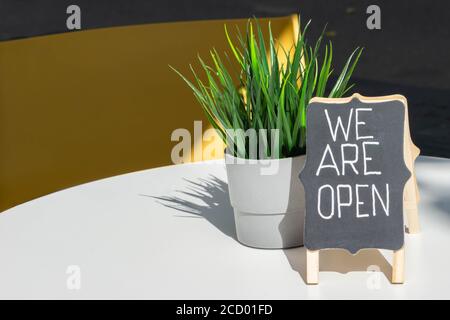 Handwritten text WE ARE OPEN and on blackboard pot with grass closeup on cafe table. Open sign as symbol of reopened small business. Open sign in cafe Stock Photo