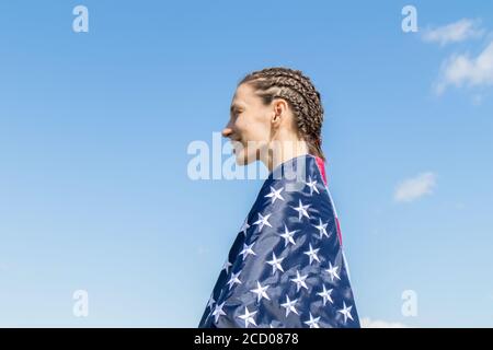 Happy american young woman with afro-braids wrapped in stripes and stars USA flag against the blue sky Stock Photo