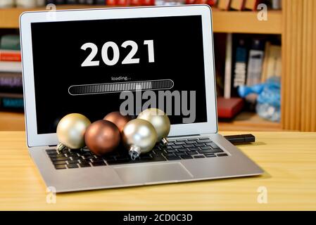on the table laptop with text - 2021 loading - on screen and with christmas decorations  on the keyboard, blurred shelf with books in the background, Stock Photo