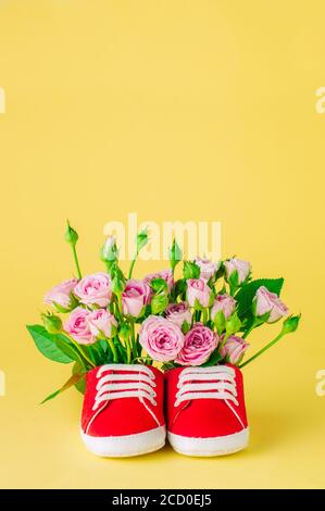 Pair of red baby shoes filled with rose flowers on yellow background. Blank space for text. Stock Photo