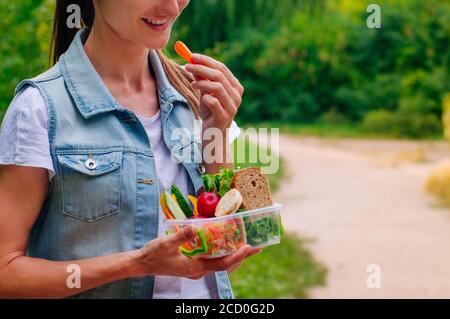 Healthy food concept: Young woman eating from lunch box filled with sandwich, crispbreads, fruits and vegetables outdoor, selective focus Stock Photo