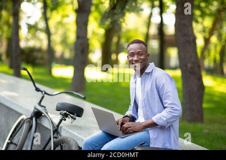 Joyful African American guy with bike working on laptop online at urban park, free space Stock Photo