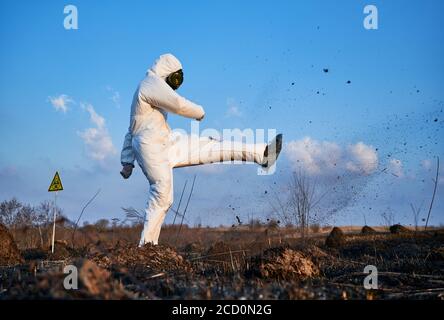 Disappointed scientist, wearing a white protective suit, gas mask and gloves, kicking soil on scorched field against blue sky. Concept of ecology and burned earth. Stock Photo