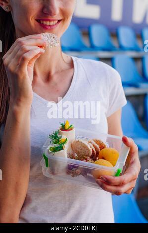 Healthy food concept: Young woman eating from lunch box filled with rolls, crispbreads, fruits and vegetables outdoor Stock Photo