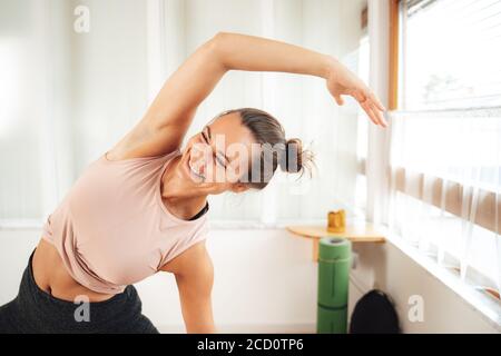 Smiling young woman in sports clothing doing side bending exercise at home Stock Photo