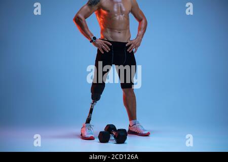 Athlete with disabilities or amputee isolated on blue studio background. Professional male sportsman with leg prosthesis training with weights in neon. Disabled sport and overcoming, wellness concept. Stock Photo