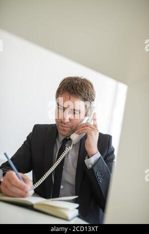 Handsome young businessman sitting at his office desk talking on white landline phone and making notes. Stock Photo