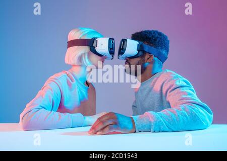 two funny people sitting in front of each other and using virtual reality goggles,playful couple in VR glasses looking at each other, close up side vi Stock Photo