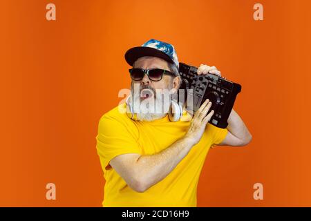 DJ-set. Portrait of senior hipster man using devices, gadgets isolated on orange studio background. Tech and joyful elderly lifestyle concept. Trendy colors, forever youth. Copyspace for your ad. Stock Photo