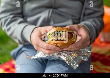 A man sits on a picnic blanket with a piece of foil on his lap, holding a hamburger in his hands; North Vancouver, British Columbia, Canada Stock Photo