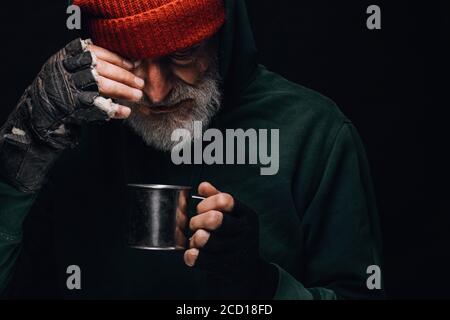Old homeless man with grey beard covering up in green decrepit clothes holding a mug of hot tea to warm himself in a cold night Stock Photo
