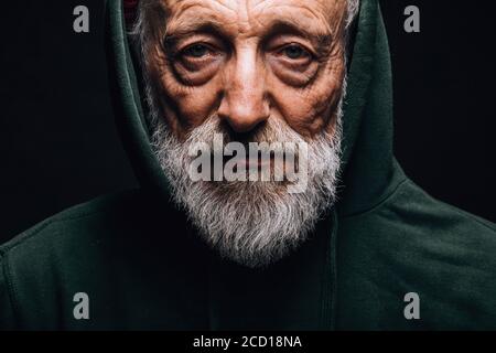 Close-up of a mature old-aged man in green hoodie with wrinkled weathered face, puffy sad eyes looking at camera with mournful expression Stock Photo