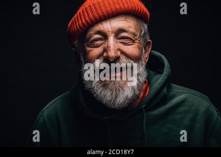 Close up portrait of happy 70-year-old optimist man with smiling wrinkled face, dressed in hipster orange hat and green hoodie, isolated over black ba Stock Photo