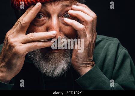 An elderly bearded man looking extremely surprised or frightened, protecting his face with hands, close up face portrait over black studio background. Stock Photo