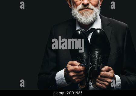 Cropped picture of unrecognizable bearded elderly man in elegant suit showing bespoke leather shoes near his chest over black background. Shoe manufac Stock Photo