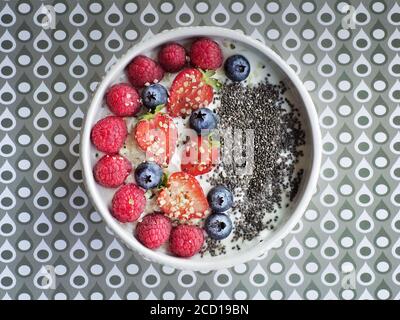 Yoghurt with berries (blueberries, strawberries and raspberries) and chia seeds in white bowl and background with pattern. Overhead shot. Stock Photo