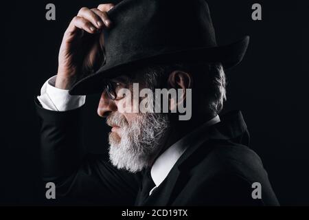 An old male actor playing an English spy role, dressed in black suit, hiding behind fedora hat, having mysterious elegant look. Isolated studio portra Stock Photo