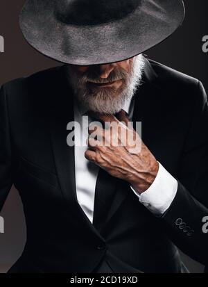 An old male actor playing an English spy role, dressed in black suit, hiding behind fedora hat, having mysterious elegant look. Isolated studio portra Stock Photo