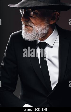An old male actor playing an English spy role, dressed in black suit, hiding behind fedora hat, having mysterious elegant look. Isolated studio portra Stock Photo