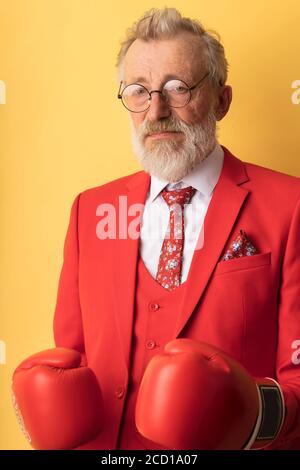 Mature business man in red formal wear wearing boxing gloves posing isolated on yellow background looking at camera with confident expression Stock Photo