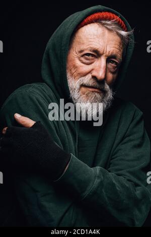 Homeless man dressed in green warm old big-sized clothing with hood on shivers from cold, tries to warm up, looking at camera with tough expression Stock Photo