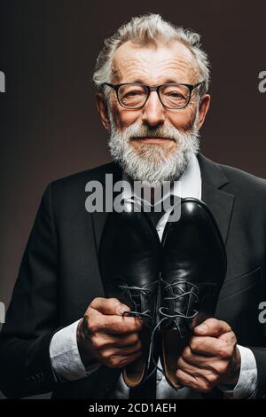 Happy elderly cobbler in elegant suit looking at camera while demonstrating bespoke leather shoes near his chest, offering to appreciate his work Stock Photo