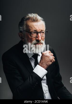 Pensive bearded grey haired stylish elderly business man in formal black suit, being deep in thoughts, keeps palms near his chin, isolated over dark Stock Photo