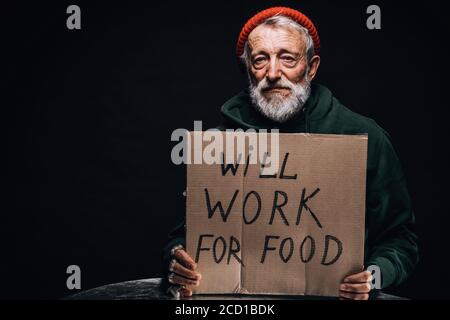 Jobless poor old aged male beggar in orange warm hat looking at camera with a handwritten sign for help holding a cup for coins in hands. Isolated stu Stock Photo