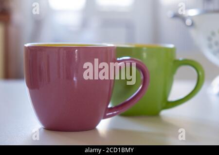 Coffee cup mockup on a kitchen table. Pink and green color blank mugs with handle for hot beverage, blurred background. Advertise, branding template Stock Photo