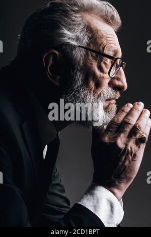Pensive bearded grey haired stylish elderly business man in formal black suit, being deep in thoughts, keeps palms near his chin, isolated over dark Stock Photo