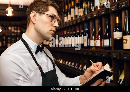 Professional male Sommelier dressed in bowtie and apron writes at notepad, putting down information on wine bottles in wine house Stock Photo