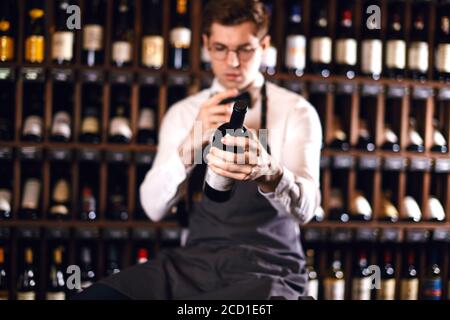 Young caucasian cavist dressed in white shirt and bowtie working in big vine shop presenting a bottle of red wine to customer Stock Photo