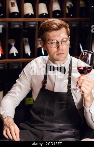 Bartender or male cavist standing near the shelves of wine bottles holds a glass of wine, looks at tint and smells flavor of wine in glass. Stock Photo