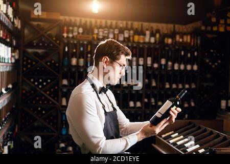 Professional male cavist examines the bottles with wine in wine shop, holding wonderful sample, ready to speak out about this wine to customers Stock Photo