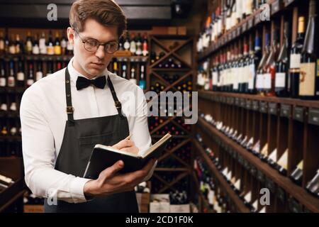 Professional male Sommelier dressed in bowtie and apron writes at notepad, putting down information on wine bottles in wine house Stock Photo