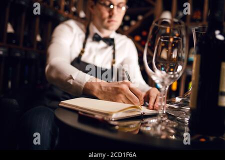 Blurred photo of male cavist making notes at his notepad about wine features and origin in wine shop, focus on clean empty wine glass Stock Photo
