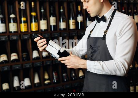 Young caucasian cavist dressed in white shirt and bowtie working in big vine shop presenting a bottle of red wine to customer Stock Photo