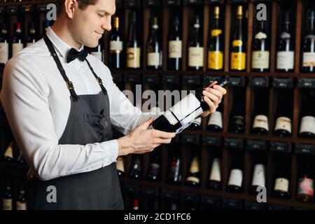 Young caucasian cavist dressed in white shirt and bowtie working in big vine shop presenting a bottle of red wine to customer Stock Photo