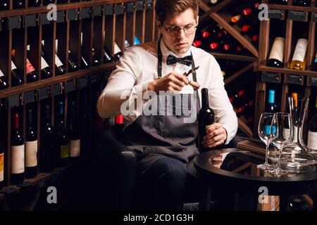 Young handsome sommelier pulling cork out of a bottle using a sommelier knife. Cropped photo of a waiter opening red wine bottle with corscrew in rest Stock Photo
