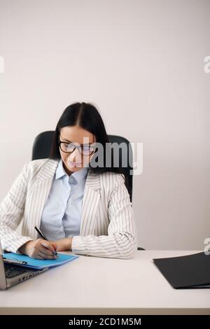 Cheerful brunette lady with important document friendly smiling and looking at camera while browsing laptop at desk in home office Stock Photo