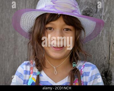 Cute little mixed-race girl (Caucasian and Southeast Asian) with long, unkempt hair wears a modern sun hat and smiles into the camera. Stock Photo