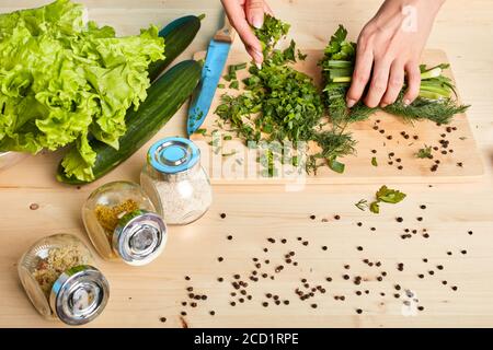 Cropped view of unrecognizable womans hands chopping scallions and making green salad with lettuce, cucumbers, parsley and different spices, cooking h Stock Photo