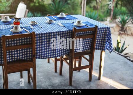 Table with empty plates and coffee cups,  blue chequered tablecloth, ready for morning breakfast, blurred green foliage in background - tropical holid Stock Photo