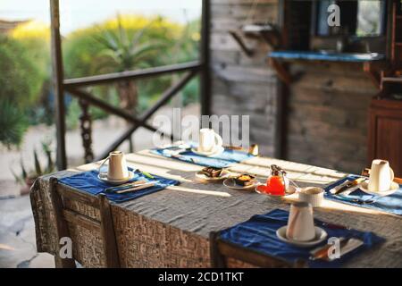 Morning sun shines on table with empty plates and coffee cups,  glass tea pot in middle, ready for morning breakfast, blurred green foliage in backgro Stock Photo