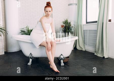 Pleased half-dressed young woman with red hair bun looks with pleasure at camera, sitting barefoot at the bathtub edge in luxurious big bathroom, prep Stock Photo