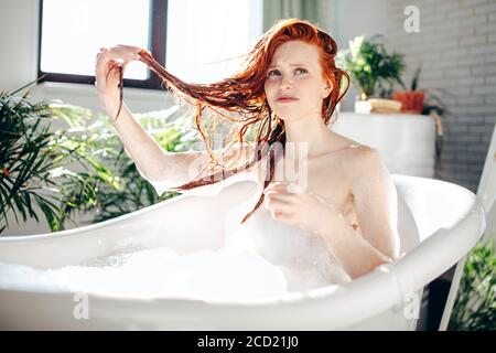 Miserable looking disappointed woman taking foam bath holding her wet red hair in hands being extremely upset with hair condition, making grimace diss Stock Photo